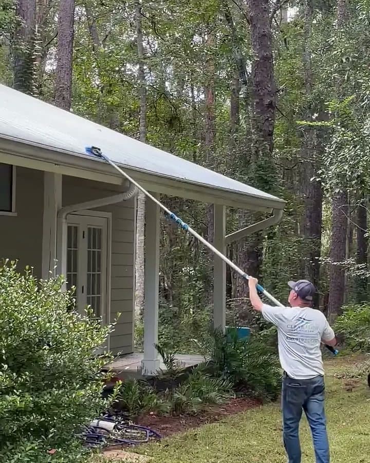 Clear Flow technician washing a house eave from the lawn with a long soft-wash pole, no ladder against the gutter